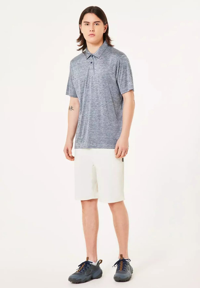 Joven de pie con camisa polo gris, pantalones cortos blancos y zapatillas oscuras, con pelo de longitud media y tatuaje en el antebrazo izquierdo.