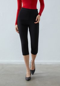 Sorte capri leggings laget av et glatt, elastisk stoff med høy midje, kombinert med en tettsittende rød langermet topp og svarte spisse hæler.
