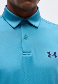 Κοντινό πλάνο ατόμου που φορά μπλε πόλο πουκάμισο Under Armour με δύο κουμπιά κουμπωμένα και εμφανές λογότυπο στο στήθος.