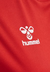 Punainen urheilupaita, joka on valmistettu teksturoidusta kankaasta. Paidassa on valkoinen "hummel"-logo ja etupuolella perhosen kaltainen kuvio.