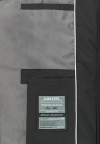 Black outerwear fabric with gray lining, featuring a rectangular label with brand details. Includes a large pocket and trim accents.