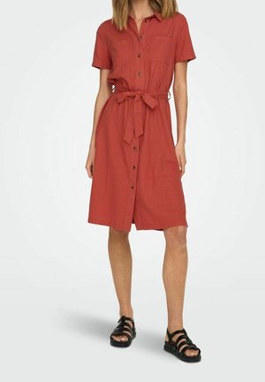 Kvinde iført en rustfarvet knapforan-kjole med korte ærmer, et bælte i taljen og sorte sandaler med stropper, stående på en hvid baggrund.