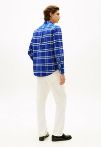 Blauwe geruite button-up blouse met witte en rode accenten, lange mouwen, achterklep, gecombineerd met witte broek en zwarte schoenen. Glenige textuur.