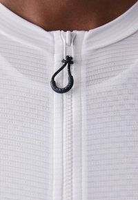 Weißes Sportoberteil mit einem Reißverschluss vorne und einem schwarzen Pull-Tab. Der Stoff hat ein strukturiertes, geripptes Muster für zusätzliche Designdetails.