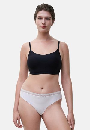 Frau trägt einen nahtlosen schwarzen BH und weiße, mit Spitze besetzte Unterwäsche, steht vor einem einfarbigen hellen Hintergrund.