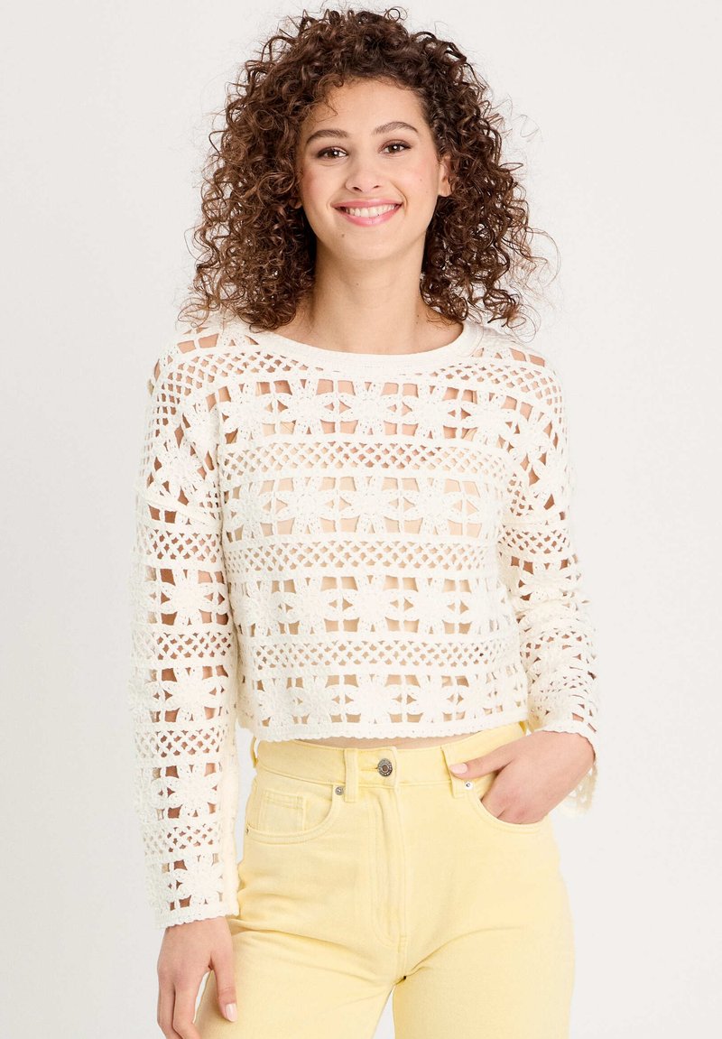 Pull en crochet crème avec un motif découpé géométrique, manches longues et design court, associé à un jean jaune taille haute.