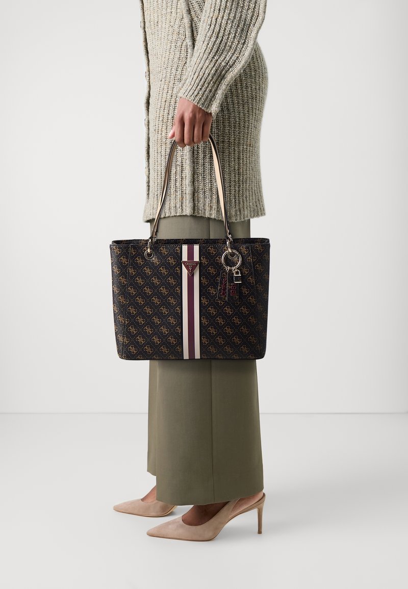 Personne portant un pull en maille beige, une jupe vert olive et des talons beige, tenant un sac à main marron à motifs avec des rayures verticales et un porte-clés.