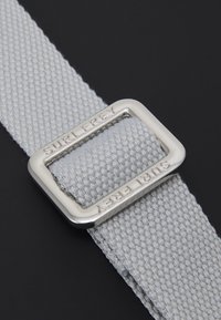 Ceinture en tissu gris clair avec une boucle en métal argenté rectangulaire gravée "SURI FREY." Motif tissé texturé sur l'ensemble de la sangle.