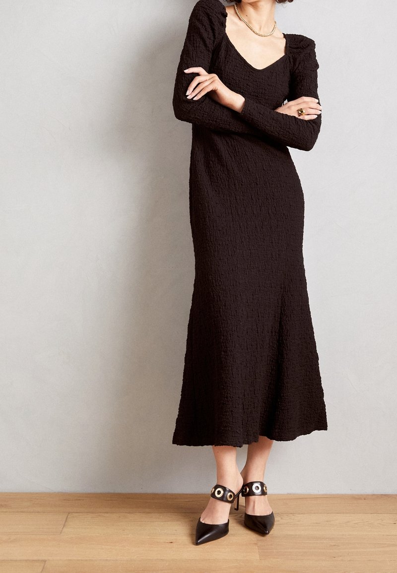 Svart, texturerad långärmad klänning med fyrkantig halsringning, kompletterad med svarta spetsiga klackskor med rundade detaljer på ankelremmen.