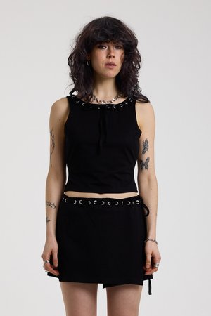 Γυναίκα με σκούρα σπαστά μαλλιά που φοράει μαύρο τοπ χωρίς μανίκια και φούστα με λεπτομέρειες από κρίκους, δείχνοντας τα τατουάζ και στα δύο χέρια της μπροστά σε απλό φόντο.