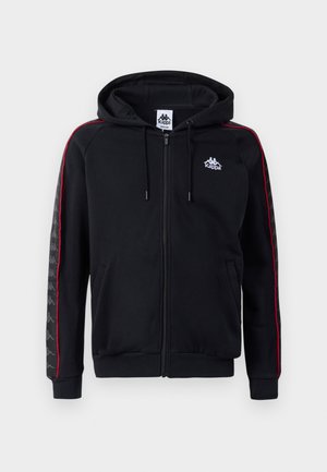 Sudadera con cremallera negra y franjas laterales rojas, capucha con cordón y logo de Kappa. Presenta patrones texturizados en las mangas y puños y dobladillo acanalados.