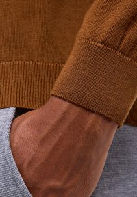 Cuff di un maglione lavorato a maglia marrone con texture a coste, che mostra una vista parziale di una mano appoggiata su una superficie di tessuto grigio.