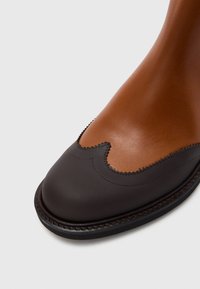 Gheata de gleznă din piele maro și maro închis, cu un design cu margine ondulată, textură netedă și cusături contrastante de-a lungul tălpii.