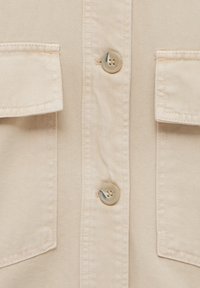 Chemise en denim beige avec deux poches poitrine, boutons de couleur contrastante et tissu à texture lisse.