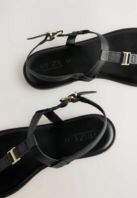 Sandalias de cuero negro con un diseño minimalista, acentuadas con una hebilla en tono dorado y tiras ajustables. Textura suave y suela plana.