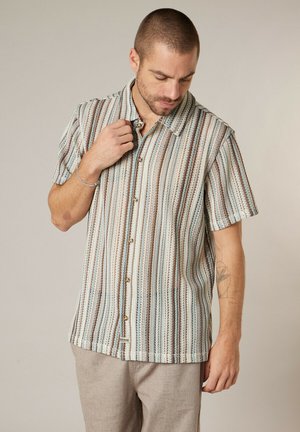 Man met kort haar die een kortärmlig, verticaal gestreept overhemd met knopen en beige broek draagt, naar beneden kijkt en de kraag aanraakt.