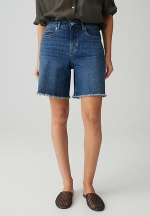 Frau trägt blaue Jeans-Knielange Shorts mit ausgefransten Säumen, ein dunkelgrünes hochgekrempeltes Hemd und schwarze gewebte Slip-On-Schuhe.