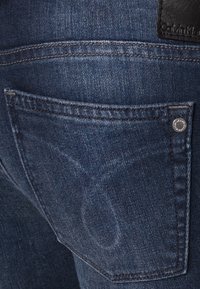Tmavě modré džíny z denimu mají zadní kapsu s malým stříbrným knoflíkem a texturovanou etiketou Calvin Klein na pase.