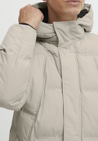 Light beige puffer jacket with a high collar, quilted texture, and a smooth finish. Features an adjustable hood and side pockets.