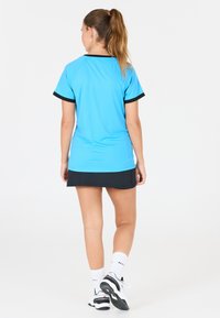 Kvinna står med ryggen vänd, iförd en ljusblå kortärmad skjorta, svart kjol, vita strumpor och vita sneakers.