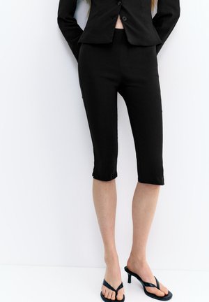 Persoon die zwarte cropped broek, zwarte blazer en zwarte sandalen met hakken draagt, staand tegen een witte achtergrond.