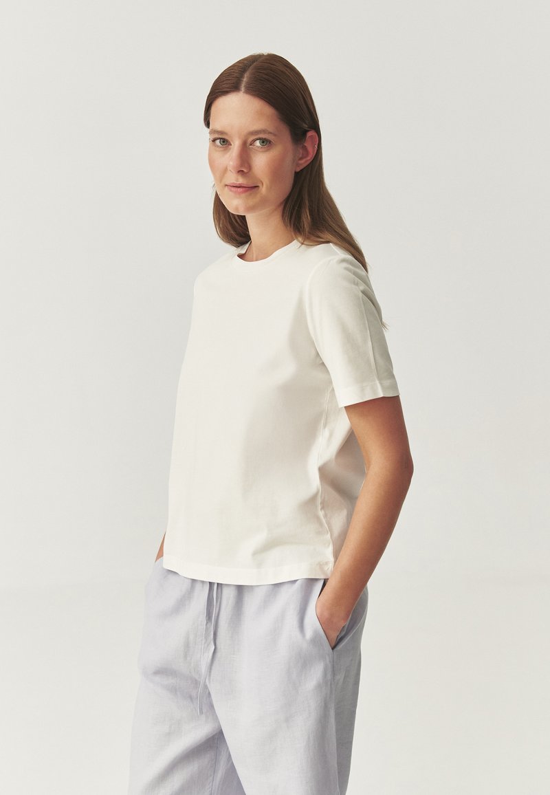 White cotton t-shirt with short sleeves and a crew neckline, featuring a relaxed fit and side slits, paired with light gray linen pants.