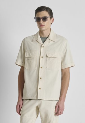 Man draagt een beige korte mouwen overhemd met borstzakken, bijpassende beige broek en donkere zonnebril tegen een effen achtergrond.