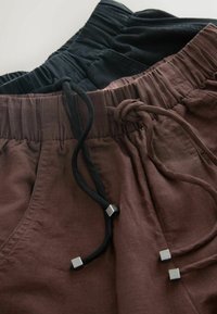 Pantalons en lin marron et noir avec tailles élastiques et cordons noirs à embouts métalliques carrés, posés en chevauchement sur une surface plane.