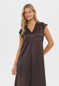 Smeđa haljina od glatkog materijala, sa V-izrezom, kratkim kap sleeves i prilagođenim gornjim dijelom te lepršavom suknjom. Minimalistički dizajn.