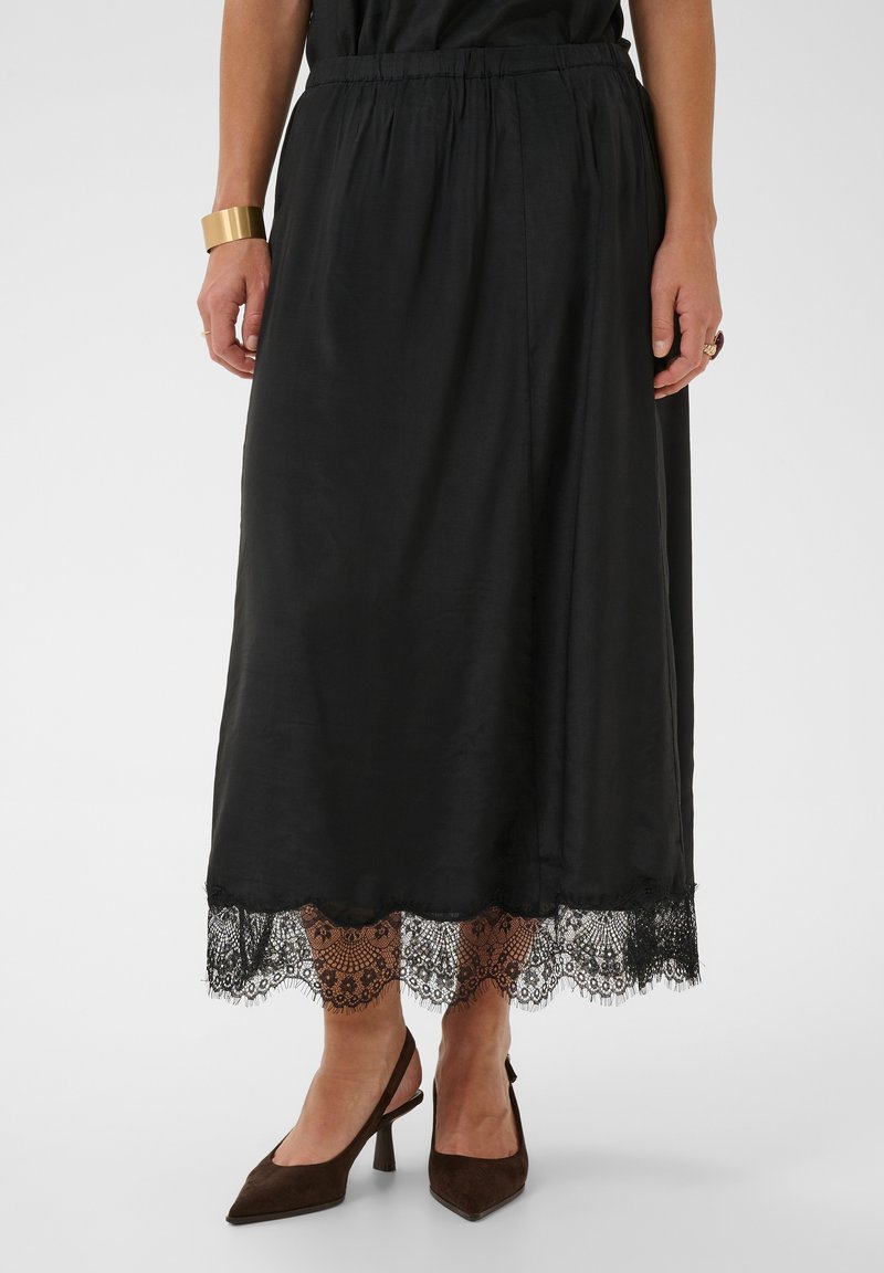 Jupe midi noire avec bordure en dentelle à l'ourlet, portée avec des talons slingback marron à bout pointu, un bracelet en or et des bagues aux mains.