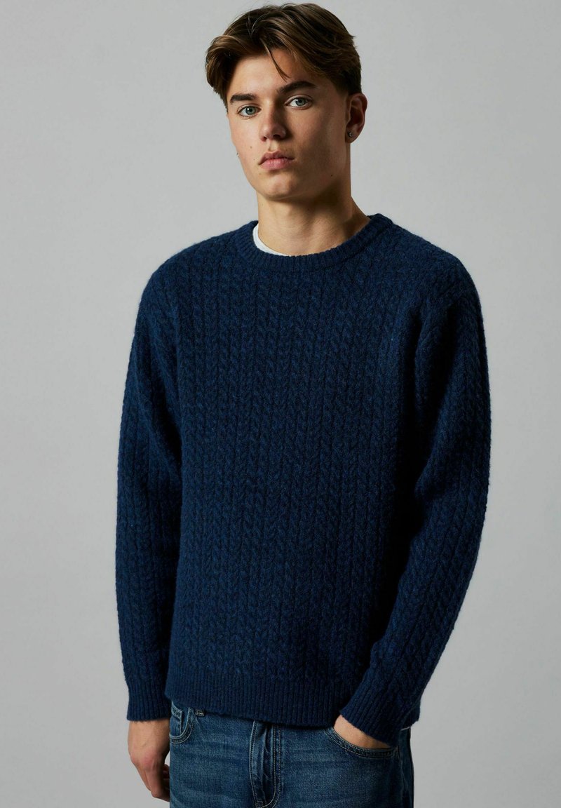Suéter de punto azul marino con un patrón de cable texturizado, cuello redondo y puños acanalados, estilizado sobre una camisa de color claro.