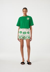 Groene katoenen t-shirt met korte mouwen en bloemenborduurwerk; witte shorts met scalloped rand en groene bloemenpatronen en decoratieve stippen. Zwarte slippers.