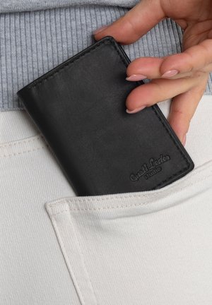 Hand placing black leather wallet labeled "Gusti Leder Studio" into white pants pocket over a gray ribbed top.