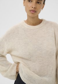 Gaiši bēšs plaša piegriezuma džemperis no mīksta dzijas auduma, ar ribotu apaļu kakla izgriezumu un smalku tekstūru. Brīvs piegriezums un gari piedurkņi.