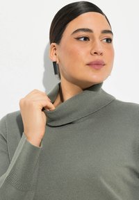 Grüner, gestrickter Rollkragenpullover mit strukturiertem Finish. Er hat einen umgeschlagenen Kragen und gerippte Manschetten und wird mit einer Hand am Hals gestylt.