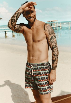 Mężczyzna bez koszulki z tatuażami tribal na całej ręce stoi na piaszczystej plaży, nosi wzorzyste kąpielówki i osłania oczy przed słońcem podnosząc rękę.