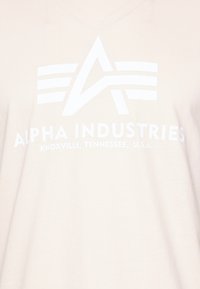 Bežs T-krekls ar baltu drukātu logotipu, kurā ir teksts "ALPHA INDUSTRIES" un simbolisks dizains ar trim horizontālām līnijām.