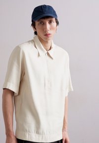 Haut blanc à manches courtes avec col et fermeture éclair à mi-longueur, associé à une casquette bleu marine. Tissu lisse, coupe décontractée et design minimaliste.