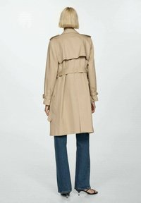Beige trench coat with a belted waist, shoulder epaulettes, and a back flap. Paired with blue wide-leg jeans and black sandals.