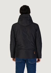 Giubbotto imbottito nero con cappuccio, dal tessuto liscio. Presenta polsini elasticizzati e un accento arancione vicino all'orlo. Indossato su jeans blu.