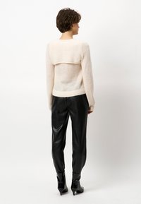 Helles, cremefarbenes Strickpullover mit einem Überwurf im Rückenbereich, kombiniert mit schwarzen Lederhosen und glänzenden Knöchelstiefeln. Weiche Textur, figurbetonter Schnitt.