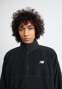 Mustal fleece-jope kõrge kraega, millel on lukk ees ja väike valge New Balance'i logo vasakul rinnal. Pehme tekstuur.