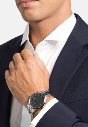 Silberne Uhr mit schwarzem Ziffernblatt, Edelstahlarmband und rundem Gehäuse. Am Handgelenk getragen, teilweise sichtbar neben einem weißen Hemd.
