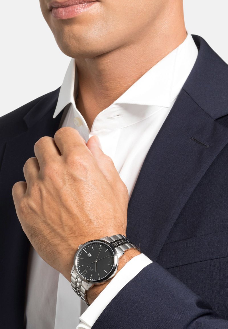 Zilveren horloge met een zwarte wijzerplaat, roestvrijstalen band en ronde kast. Dragen om een pols, gedeeltelijk zichtbaar naast een wit overhemd.