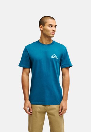 Man draagt een blauw T-shirt met korte mouwen met een klein wit logo op de linkerborst en beige broek, kijkt naar rechts.