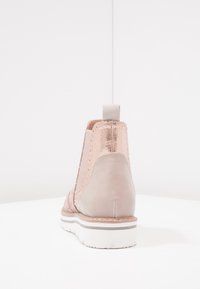 Jednoduchá růžová kotníková bota s bílou vroubkovanou podrážkou, kovovým růžově zlatým kloubem na patě a poutkem, zobrazená z pohledu zezadu na bílé ploše.