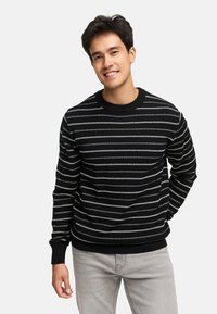 Sort og hvidstribet strikket sweater med rund halsudskæring, lange ærmer og ribbede manchetter. Båret over lysegrå jeans.