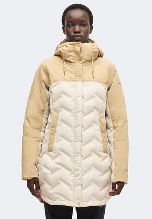 Kvinde iført en beige og lys tan quiltet vinterjakke med hætte, der står oprejst mod en ensfarvet hvid baggrund.