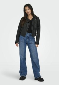Veste de biker en cuir noir avec des zippers argentés, associée à un jean large bleu taille haute. Le mannequin se tient devant un fond uni.