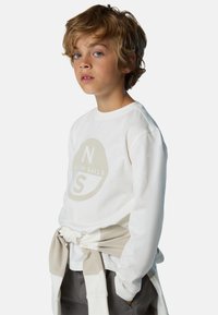 Maglietta bianca a maniche lunghe con una grafica circolare beige contenente il testo "N S". Abbinata a pantaloni grigi e un cardigan beige annodato in vita.